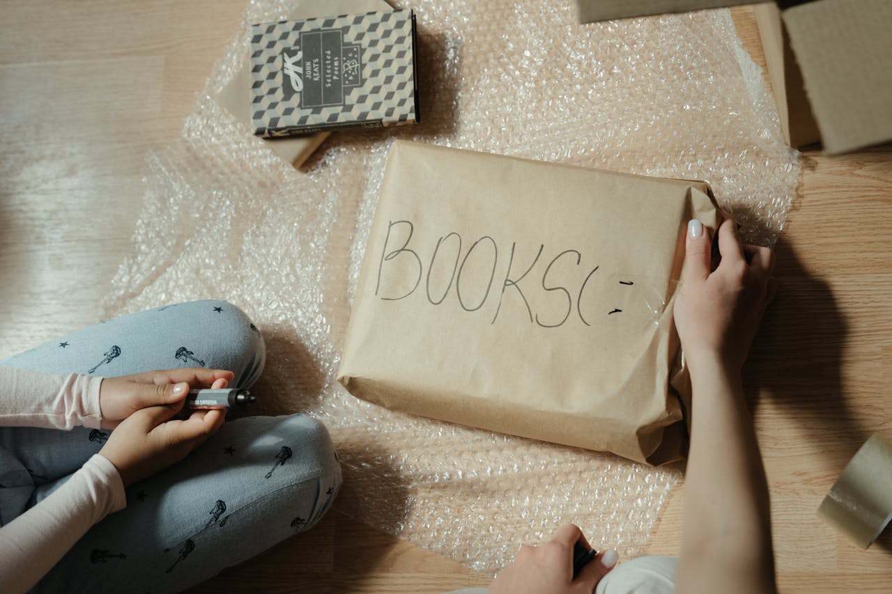 Two people carefully pack books using bubble wrap and brown paper for safe moving.